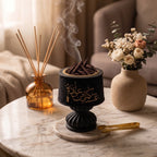 A black incense burner with gold Arabic calligraphy sits on a marble coaster on a round marble table, with incense sticks smoking upward. A reed diffuser bottle and a vase of dried flowers are styled nearby in a warm, softly lit interior setting.